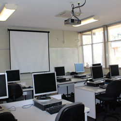 There are two rows of desks set up facing the front of the room. Two computers are at each desk. The front of the room has a whiteboard and a projector set up