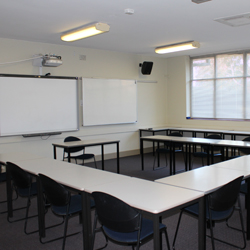 There are desks set up in a u-shape facing the front of the room. The front of the room has a whiteboard and a projector set up.