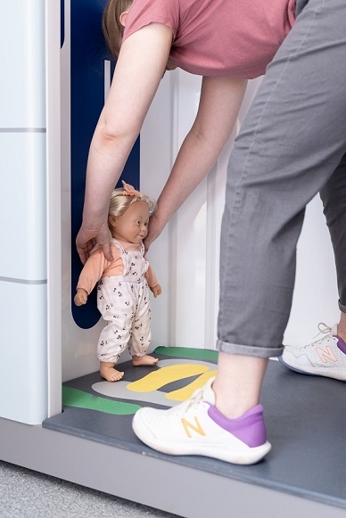 A woman stands outside the scanner, hands on the shoulders of a child who is standing legs apart and arms by their sides in the scanner.