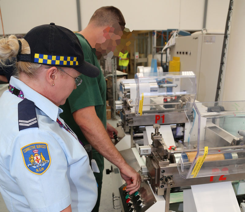 A person in a blue Correctional Officer uniform watches as a person in a green T-shirt operates a machine on a table.