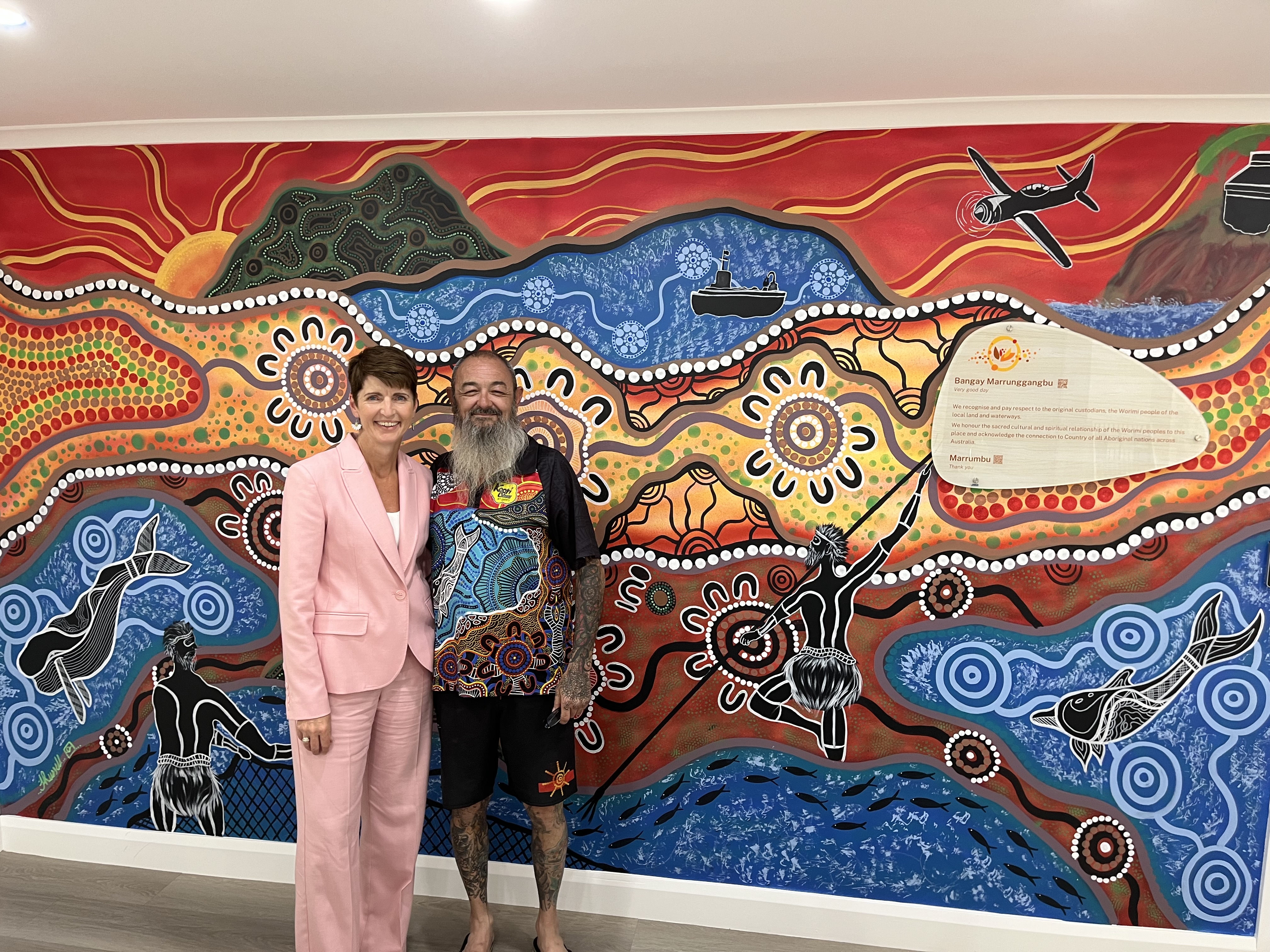 A traditional Aboriginal artwork with earth based and ocean based colours. Minister Kate Washington is wearing a pink suit and The artist Jason Russell is wearing a tshirt and shorts 
