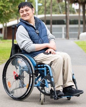 Quang Nguyen is setting in a wheelchair in a park setting. He is wearing a blue vest with a pale blue shirt underneath and bone coloured long pants. He has short black hair