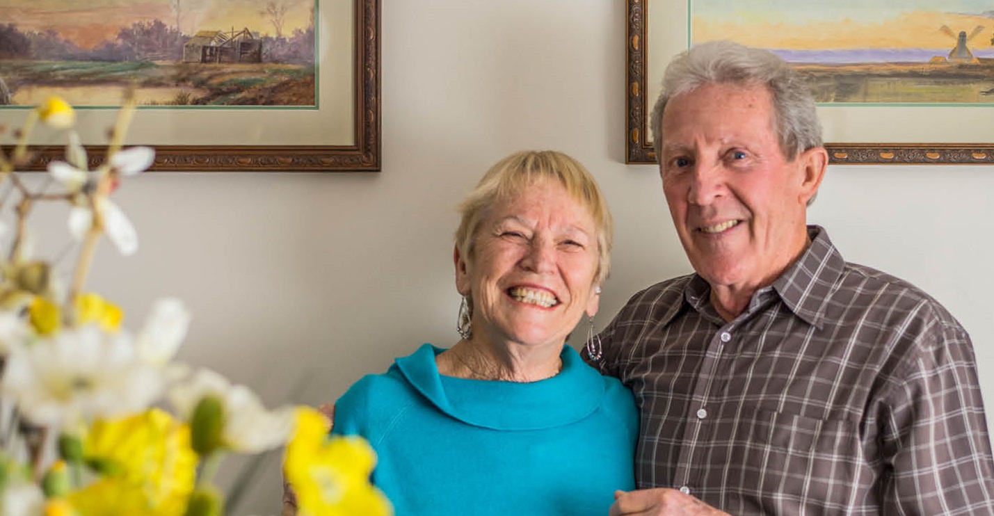 Two elderly individuals standing next to paintings, smiling.