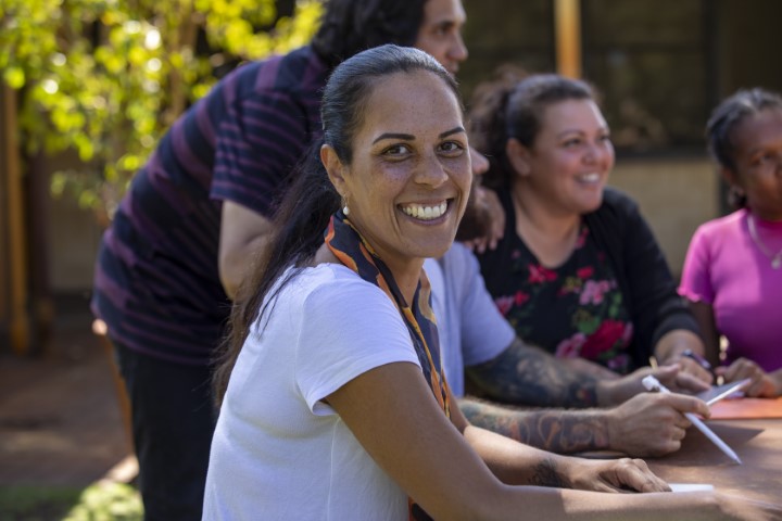 A group of Aboriginal community workers meeting at an outdoor table. A woman in the foreground is smiling at the camera.